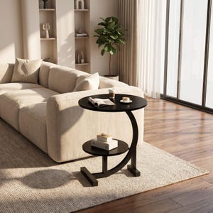 May include: A black C-shaped side table with a circular top and a smaller circular lower shelf. The table features a curved support frame and a flat base. On the top surface, there is an open book and a cup. Two stacked books sit on the lower shelf.
