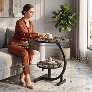 May include: A black C-shaped side table with two round shelves featuring a floral pattern. The top shelf is 11.81 inches in diameter, the bottom shelf is 9.84 inches in diameter, and the overall height of the table is 25.6 inches.