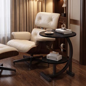 May include: A round black side table with a C-shaped frame and a lower shelf. The table features a dark marble-like surface holding a cup and books, with a white tissue box on the lower shelf, positioned next to a cream-colored lounge chair.