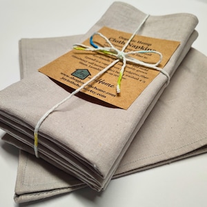 May include: A bundle of beige cloth napkins tied with white string and a brown tag. The tag reads "Organic Hemp Cloth Napkins" and includes website details. The napkins are made of a natural fibre and are absorbent.