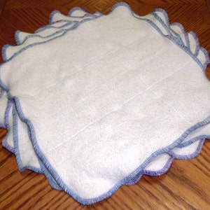 May include: A stack of white cotton cloths with blue and purple stitching around the edges. The cloths are folded and stacked on top of each other.