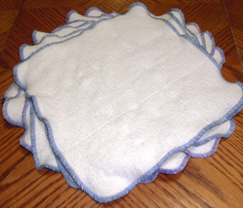 May include: A stack of white cotton cloths with blue and purple stitching around the edges. The cloths are folded and stacked on top of each other.