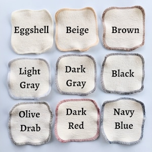 May include: Nine fabric swatches in various colours, including eggshell, beige, brown, light grey, dark grey, black, olive drab, dark red, and navy blue. Each swatch is a square with a contrasting trim around the edges.