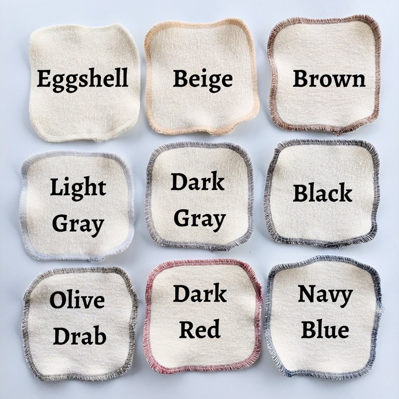 May include: Nine fabric swatches in various colours, including eggshell, beige, brown, light grey, dark grey, black, olive drab, dark red, and navy blue. Each swatch is a square with a contrasting trim around the edges.