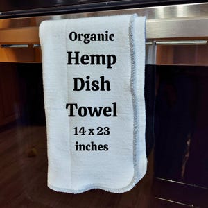 May include: A white organic hemp dish towel with black text that reads "Organic Hemp Dish Towel 14 x 23 inches". The towel is hanging on a silver oven door.