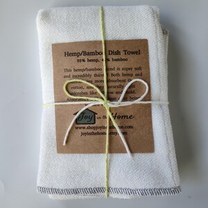 May include: A set of four white hemp and bamboo dish towels tied with a green and white twine bow. The towels have a grey trim and are packaged with a brown label that reads "Hemp/Bamboo Dish Towel 55% hemp, 45% bamboo" and includes information about the product's absorbency and antimicrobial properties. The label also includes the company name "Joy in the Home" and website information.