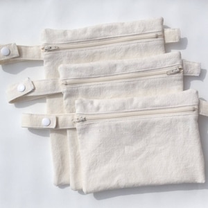 May include: Three natural-colored reusable cloth bags with zipper closures and snap closures on the handles. The bags are stacked on top of each other.