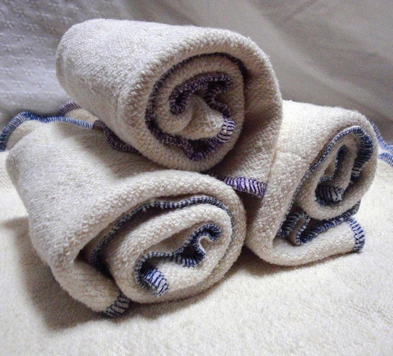 May include: Three white cotton cloth diapers rolled up with blue and purple trim. The diapers are folded and stacked on top of each other.