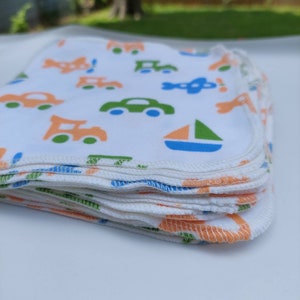 May include: A stack of white cloth burp cloths with a colourful print of cars, trucks, and sailboats. The cloths have a light blue, green, and orange trim.