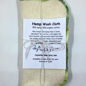 Peut inclure: Un torchon en tissu de chanvre pli&eacute; avec une bordure verte, attach&eacute; avec de la ficelle. L'&eacute;tiquette indique "Hemp Wash Cloth 55% hemp 45% organic cotton" et explique les avantages du chanvre. Le texte comprend &eacute;galement le nom de la marque "Joy in the home" et l'adresse du site Web "Joyinthehome.etsy.com".