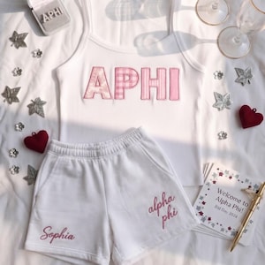 May include: A white ribbed tank top with pink patterned letters spelling APHI, paired with white shorts featuring the embroidered names Sophia and alpha phi in pink. A small box with APHI text and a card reading Welcome to Alpha Phi! Bid Day 2024 are included.