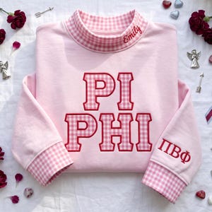 May include: A light pink sweatshirt featuring a mock neck collar and sleeve cuffs in a pink and white gingham pattern. The front displays the text PI PHI in large gingham-filled letters with red outlines. The name Emily is embroidered on the collar.