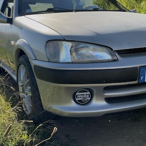 Könnte beinhalten: Ein silberner Peugeot-Kleinwagen mit einer vorderen Stoßstange mit schwarzer Zierleiste, klaren Scheinwerfern und einem runden Hella-Nebelscheinwerfer. Die Windschutzscheibe zeigt oben den Markennamen Peugeot, und das Auto steht auf einem Feldweg.