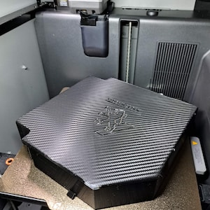 Könnte beinhalten: Eine schwarze, 3D-gedruckte hexagonale Abdeckung mit einer Kohlefaser-Textur auf der Oberseite. In der Mitte befindet sich ein geprägtes Peugeot-Löwenlogo sowie der Markenname. Das Objekt befindet sich auf einem strukturierten Metalldruckbett.