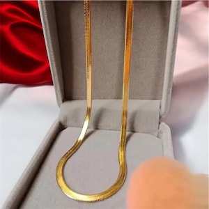 Op de afbeelding: Een goudkleurige visgraatketting met een plat, flexibel en glad schakelontwerp. De ketting wordt gepresenteerd in een met lichtgrijs fluweel bekleed juwelendoosje, waarbij het reflecterende metalen oppervlak en het patroon zichtbaar zijn.