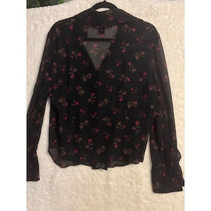 May include: A long-sleeved black blouse made of a sheer, lightweight fabric featuring a repeating pattern of small red roses with green stems and leaves. The garment has a collared neckline, a button-down front, and cuffed sleeves with a loose fit.