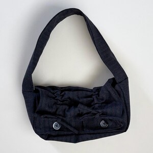 May include: A dark charcoal grey shoulder bag featuring a single wide strap, a gathered fabric design across the front, and two small exterior pockets each secured with a single matching button. The bag is constructed from a textured fabric.