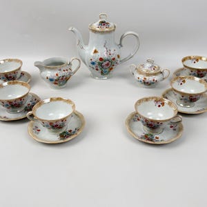Może przedstawiać: Zestaw do herbaty z porcelany zawierający dzbanek z pokrywką, mlecznik, cukiernicę z pokrywką oraz pięć filiżanek ze spodkami. Każdy element jest biały, ozdobiony kolorowym wzorem kwiatowym oraz złotymi wykończeniami na brzegach i uszkach.