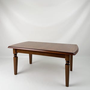 Peut inclure: Une table basse rectangulaire en bois avec une finition brun riche et un grain de bois visible. La table présente un plateau lisse et plat aux coins légèrement arrondis et quatre pieds fuselés robustes avec un détail arrondi décoratif.