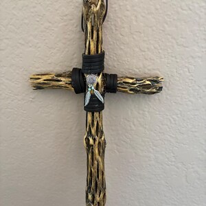 May include: A wall-hanging cross crafted from textured, porous cholla wood with a tan and dark brown finish. The center is wrapped in black leather cord and features a silver-toned circular charm with two small dangling feather pendants.