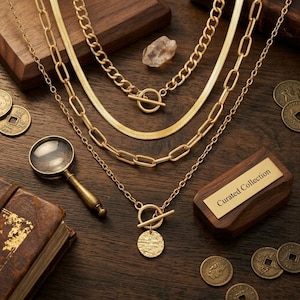 May include: A layered gold-tone necklace set featuring a thick chain, a flat herringbone chain, and a chain with a toggle clasp and a textured circular pendant. The set is displayed with a magnifying glass, coins, and a wooden box labeled Curated Collection.