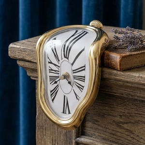May include: A gold-toned melting-style analog clock with a white face and black Roman numerals. The clock features a curved, distorted frame that allows it to drape over the edge of a surface. It has two black hands and a central winding knob.
