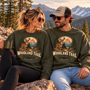 May include: Two olive green crewneck sweatshirts featuring a circular graphic of a deer in a forest with mountains and a sunset. The text reads Woodland Trail and Explore the Wild. The design includes pine trees, rocks, and foliage elements.