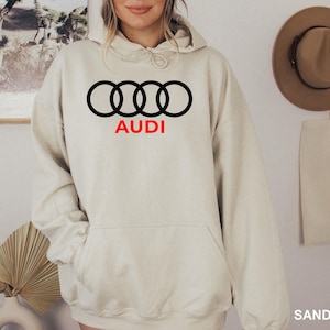 May include: A sand-colored hooded sweatshirt featuring the black Audi four-ring logo centered on the chest with the word AUDI printed in red underneath. The garment includes a front kangaroo pocket, long sleeves, and a drawstring hood.