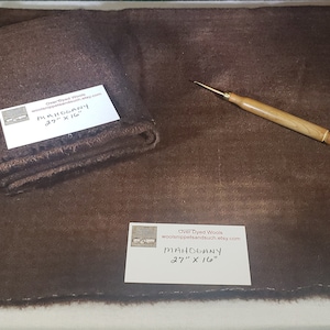 May include: A mahogany-colored wool cloth, partially rolled, with a needle tool. The cloth measures 27 x 16 inches. A white label with text is visible. The overall theme is craft supplies.