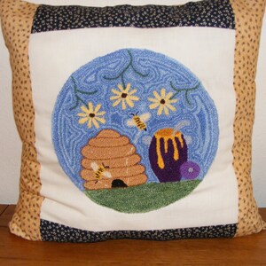 May include: A decorative pillow with a central embroidered design featuring a beehive, honey pot, and bees within a blue circle. The pillow has a white center, bordered by tan and navy blue patterned fabric.