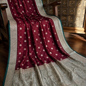 May include: A deep maroon fabric featuring a repeating pattern of small gold circular motifs. The fabric has a wide, ornate border in metallic gold and dark teal with intricate floral detailing and a finished edge with dark red tassels.