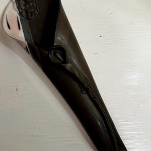 May include: A wall-mounted ceramic vase shaped like a long, tapered cone. The exterior is a matte dark brown with a sculpted floral stem and bud detail. The interior opening reveals a smooth, cream-colored surface with subtle pink and black accents.
