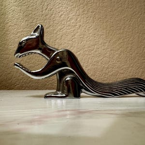May include: A shiny, silver-toned metal nutcracker shaped like a stylized squirrel. The object features a hinged jaw mechanism, a curved tail with etched line details, and a smooth, reflective surface finish throughout its entire body structure.