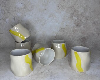 May include: A collection of six off-white ceramic cups with irregular, organic shapes. Each cup features a single, hand-painted vertical yellow brushstroke accent. The cups are arranged in a cluster, with some standing upright and others tilted.