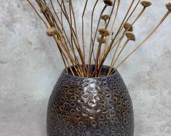 May include: A rounded ceramic vase with a dark brown and blue textured surface featuring a repeating pebble-like pattern. The vase is filled with a collection of thin, dried brown stems topped with small, round, fuzzy seed heads.