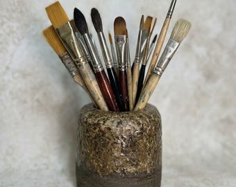 May include: A collection of various paintbrushes with wooden and black handles and metal ferrules, held upright in a cylindrical ceramic container. The container features a textured, mottled brown and tan glaze on the top and a dark brown base.