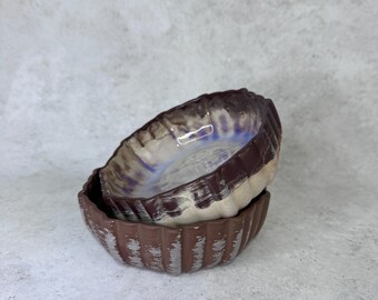 May include: Two small, round ceramic bowls stacked together. The bowls feature a dark brown exterior with vertical fluted ridges and a lighter, iridescent, pearlescent glaze on the interior that shifts between white, blue, and purple tones.