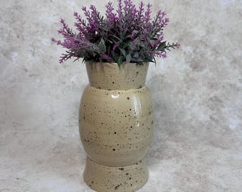 May include: A ceramic vase with a glossy cream-colored glaze and dark brown speckles throughout. The vase has a rounded body, a flared base, and a slightly tapered neck. It is filled with a cluster of artificial purple lavender-like flowers.