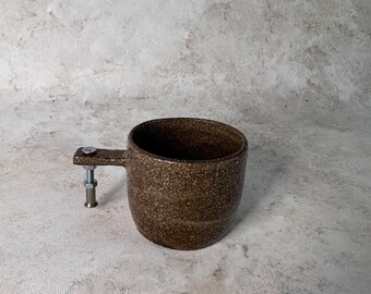 May include: A brown ceramic mug with a speckled texture and a unique handle constructed from a metal bolt, a nut, and a cylindrical metal spacer. The mug has a cylindrical body with a wide opening and a dark interior, showcasing a contrast between the earthy clay and the industrial hardware components.