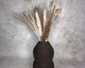 May include: A dark brown, textured, double-bulb shaped vase containing an arrangement of dried grasses, including fluffy white pampas grass plumes and slender, tan-colored bunny tail stems that extend upwards from the narrow neck of the vessel.