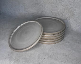 May include: A set of six round ceramic plates with a smooth, light gray finish and a subtle, swirling texture on the surface. One plate is positioned upright in the foreground, while the remaining five plates are stacked neatly behind it.