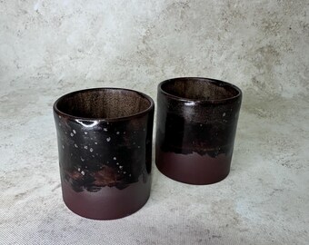 May include: Two cylindrical ceramic cups featuring a two-tone design. The upper portion of each cup has a glossy, dark brown to black glaze with subtle white speckles, while the lower portion features a matte, solid dark maroon finish.