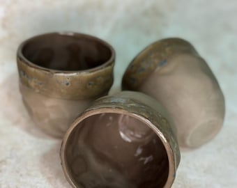 May include: Three ceramic cups with a matte beige lower body and a glossy, textured brown rim. The cups have a rounded, organic shape with a smooth interior finish. The rims feature subtle blue and gold speckles within the brown glaze.