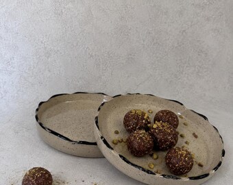 May include: Two round, speckled ceramic plates with irregular, dark-rimmed edges. One plate holds five dark brown, spherical food items topped with crushed green nuts, while a single identical food item sits separately on the surface nearby.