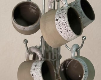 May include: A metal mug tree stand with a wooden base holds four ceramic mugs. Each mug features a matte tan body with a speckled white and grey glaze around the rim and handle. The mugs are arranged on curved hooks around the central metal pole.