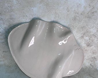 May include: A light beige ceramic dish with a smooth, glossy finish and a wavy, organic rim. The surface features subtle speckles throughout the material, and the overall shape is asymmetrical with deep, flowing indentations along the edges.