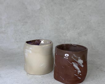 May include: Two small ceramic cups with irregular, organic shapes. One cup is a light cream color with a smooth finish, while the other is a deep reddish-brown with white marbled streaks and a glossy, reflective surface on the upper rim.