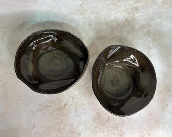 May include: Two small, round, dark brown ceramic bowls with a glossy, reflective interior finish and a textured, matte exterior rim. The bowls feature irregular, organic shapes with slightly uneven edges, showcasing a deep, dark glaze.