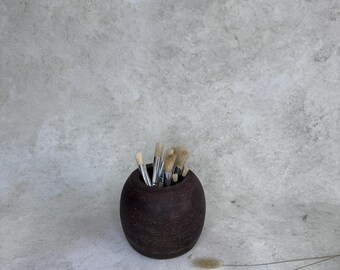May include: A dark brown, spherical container with a textured surface holding a collection of artist paintbrushes with silver ferrules and light-colored bristles. The container has a circular opening at the top for storing the brushes.