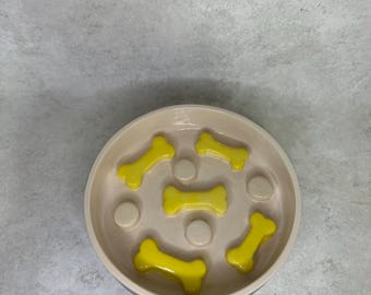 May include: A round, beige slow-feeder bowl for pets featuring a series of raised, bright yellow bone-shaped obstacles and small circular bumps integrated into the base to help regulate the speed of food consumption during mealtime.